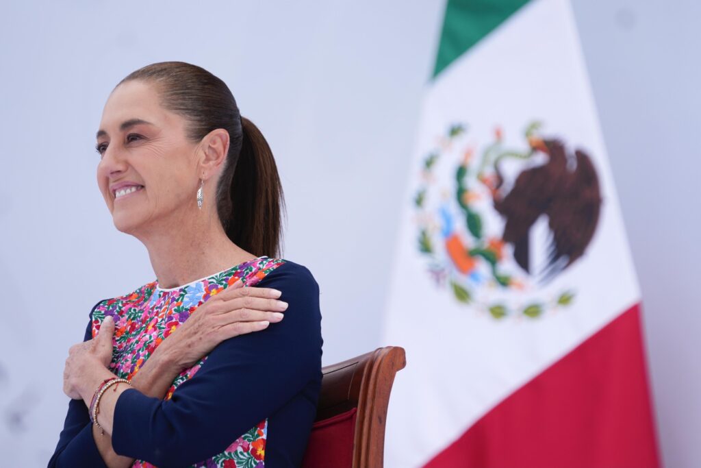 Claudia Sheinbaum sonríe sentada junto a la bandera de México durante el acto.